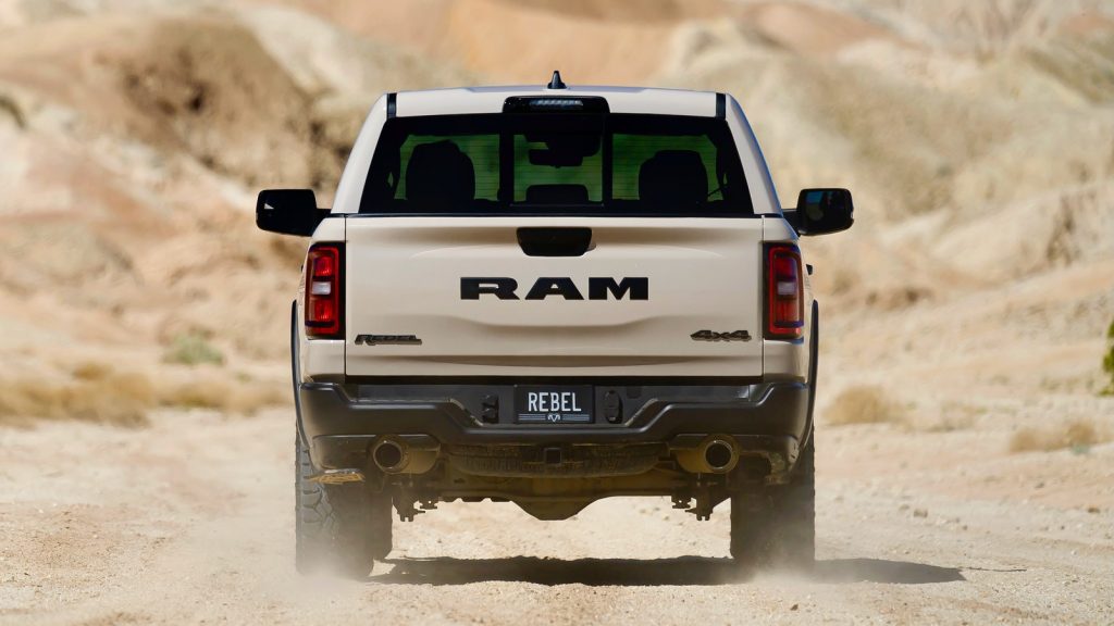 Rear view of a Ram 1500 Rebel pickup truck driving through a desert trail with bold RAM lettering on the tailgate.