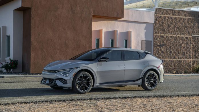 2026-kia-ev6-pricing-sales-decline-featured Silver 2026 Kia EV6 electric crossover parked in profile against modern brown architectural backdrop with geometric building…