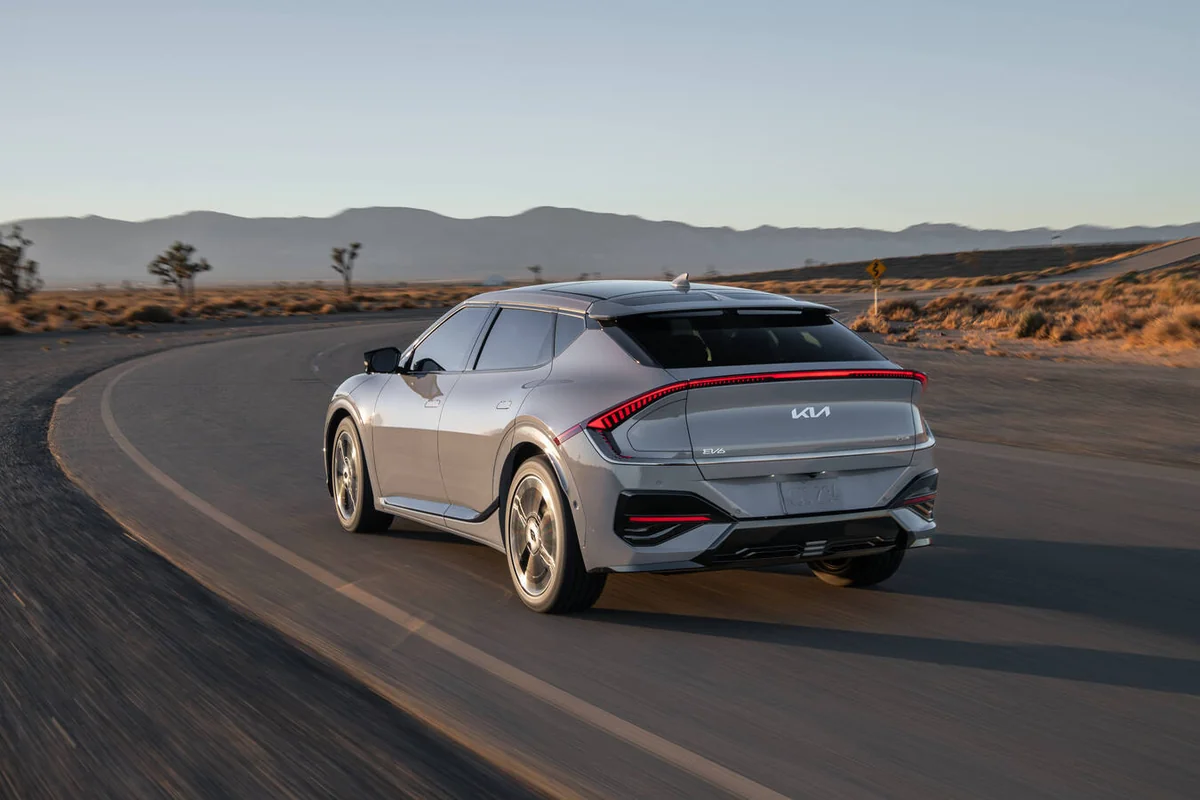 Silver 2026 Kia EV6 electric vehicle driving on desert road with mountains in background, showing rear three-quarter view wi…