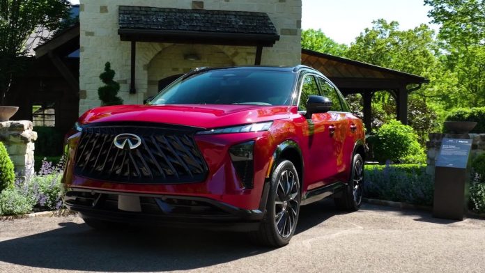 2027-infiniti-qx65-pricing-launch-featured Red 2027 Infiniti QX65 luxury SUV parked in front of stone house with landscaped grounds, side three-quarter view showcasing…