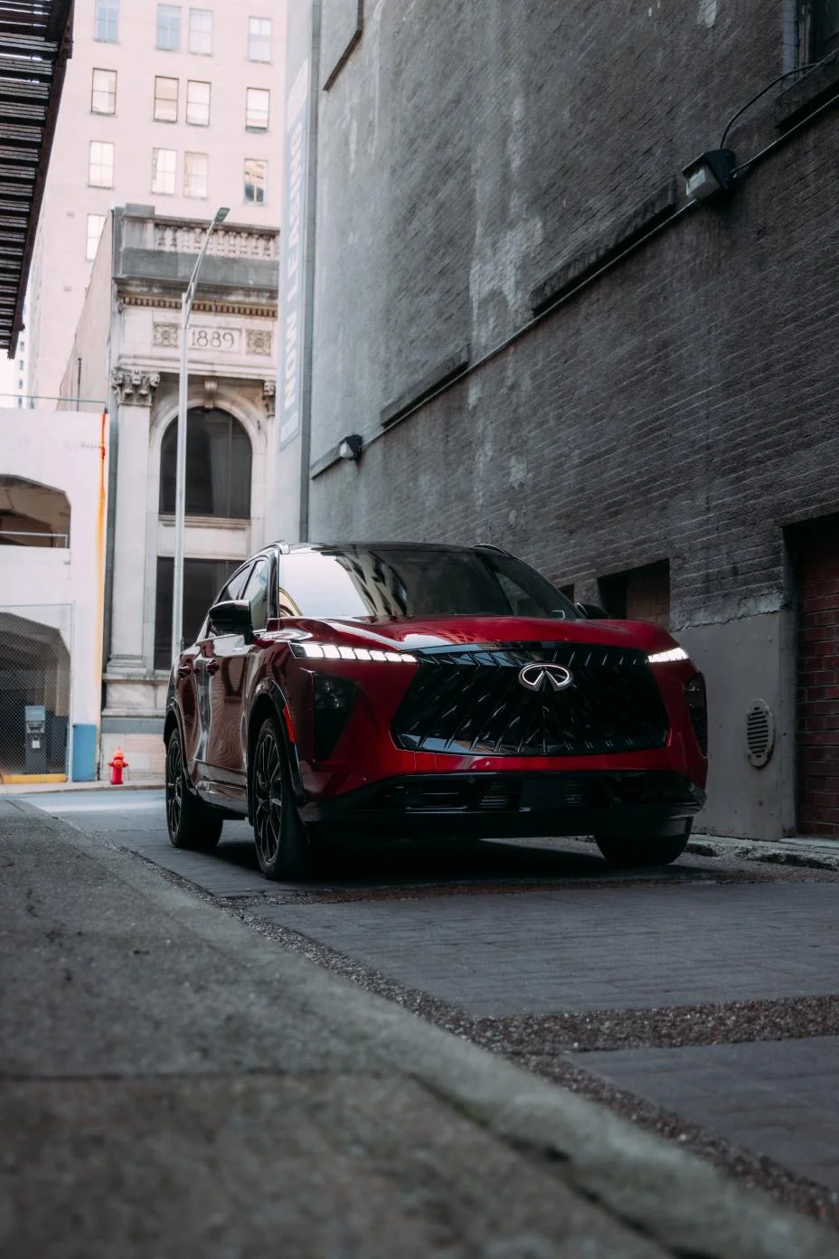 Deep red 2027 Infiniti QX65 parked in urban alley between brick buildings, front three-quarter view highlighting bold grille…
