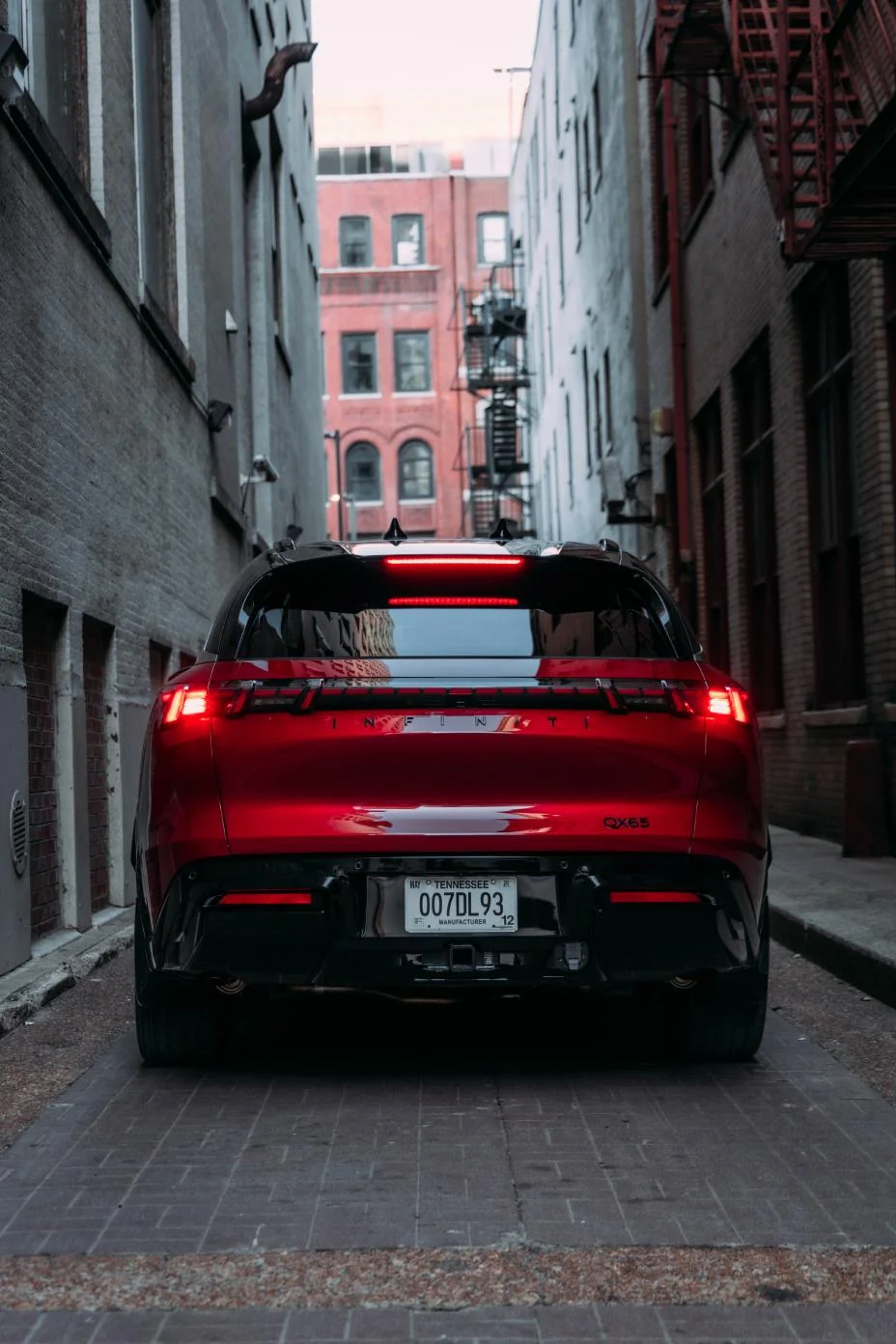Red 2027 Infiniti QX65 viewed from rear in narrow urban alley with brick buildings, illuminated taillights visible.