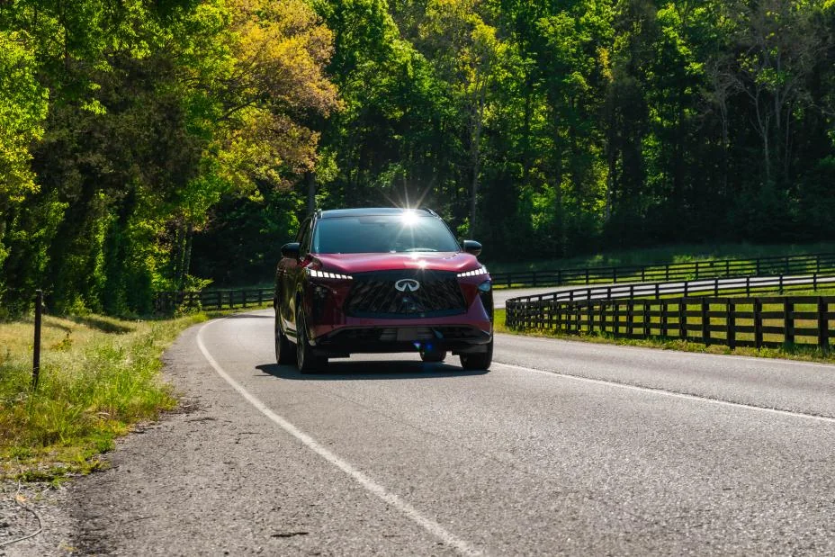 Deep crimson 2027 Infiniti QX65 driving on tree-lined country road with white fence, front three-quarter view highlighting l…