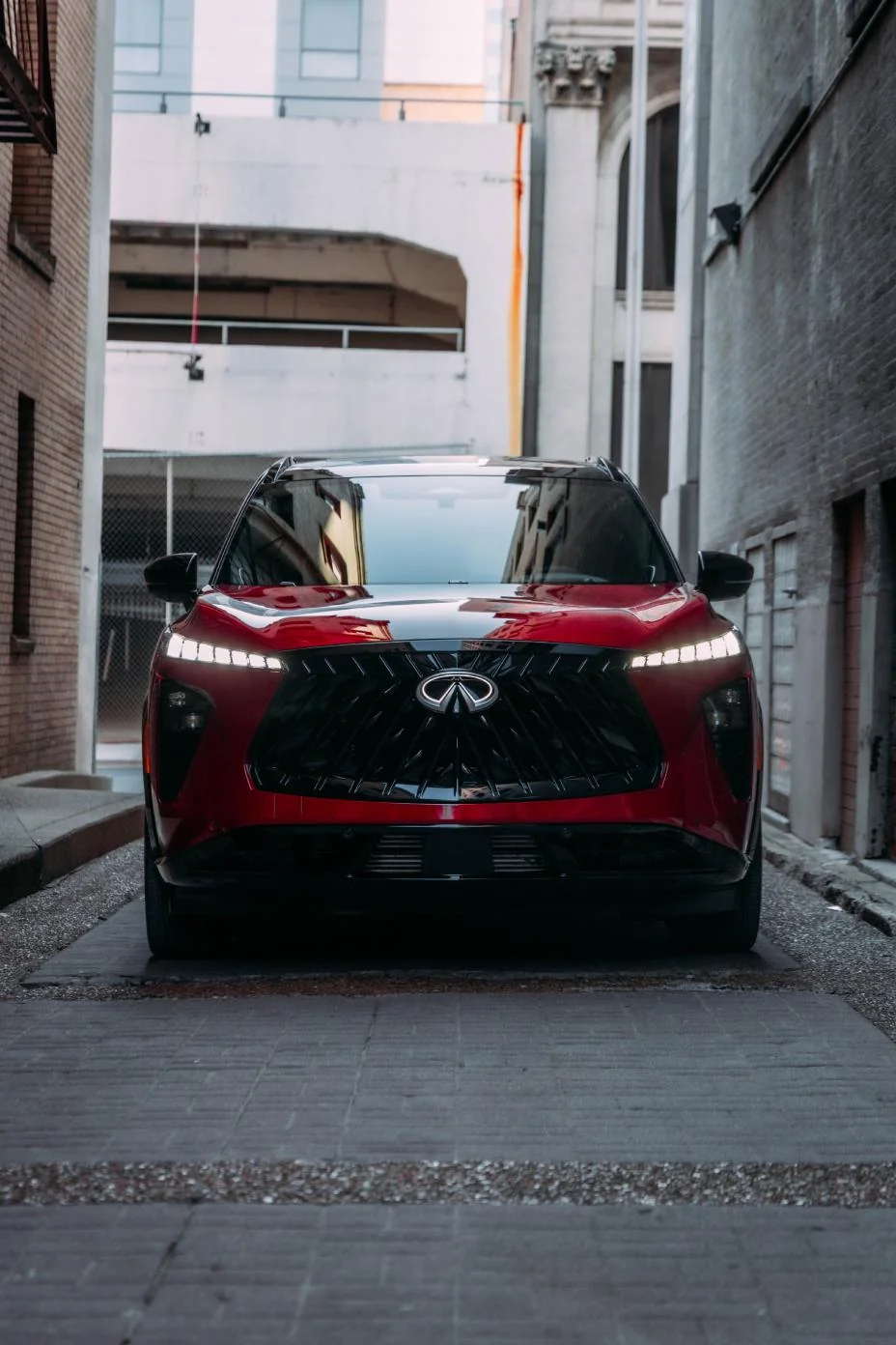 Red 2027 Infiniti QX65 parked in urban alley, front view showing illuminated grille and headlights between brick buildings.
