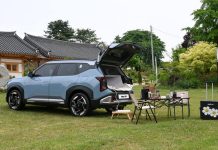 2027 Kia Seltos Grows Larger, Adds Hybrid, Targets 189 Countries Light blue 2027 Kia Seltos with open tailgate parked on grass next to camping setup and traditional Asian architecture.