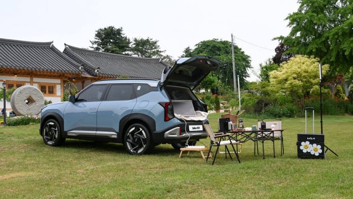 Light blue 2027 Kia Seltos with open tailgate parked on grass next to camping setup and traditional Asian architecture.