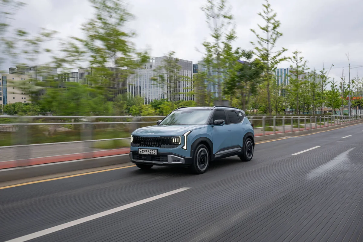 Blue 2027 Kia Seltos compact SUV driving on urban highway with modern waterfront buildings and trees blurred in background