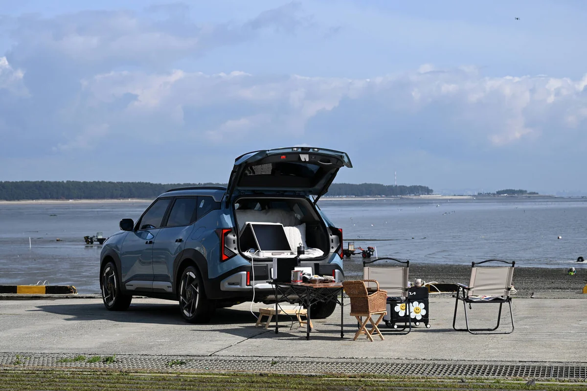 Blue 2027 Kia Seltos with open tailgate displaying camping gear and chairs at a beach parking lot overlooking the water.