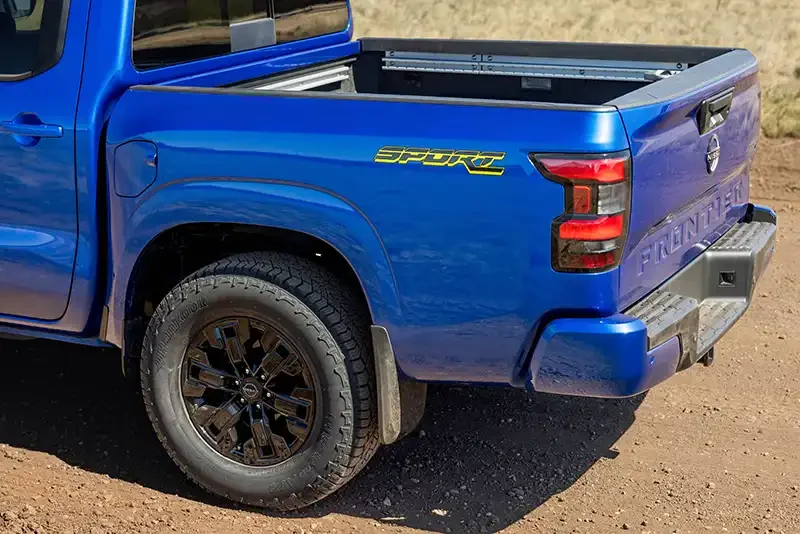 Blue 2027 Nissan Frontier Sport Edition with open bed, viewed from rear quarter angle showing all-terrain tires and Sport ba…