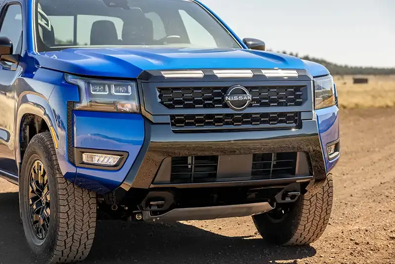 Bright blue 2027 Nissan Frontier with aggressive front grille and LED headlights parked on dirt terrain, showcasing rugged a…