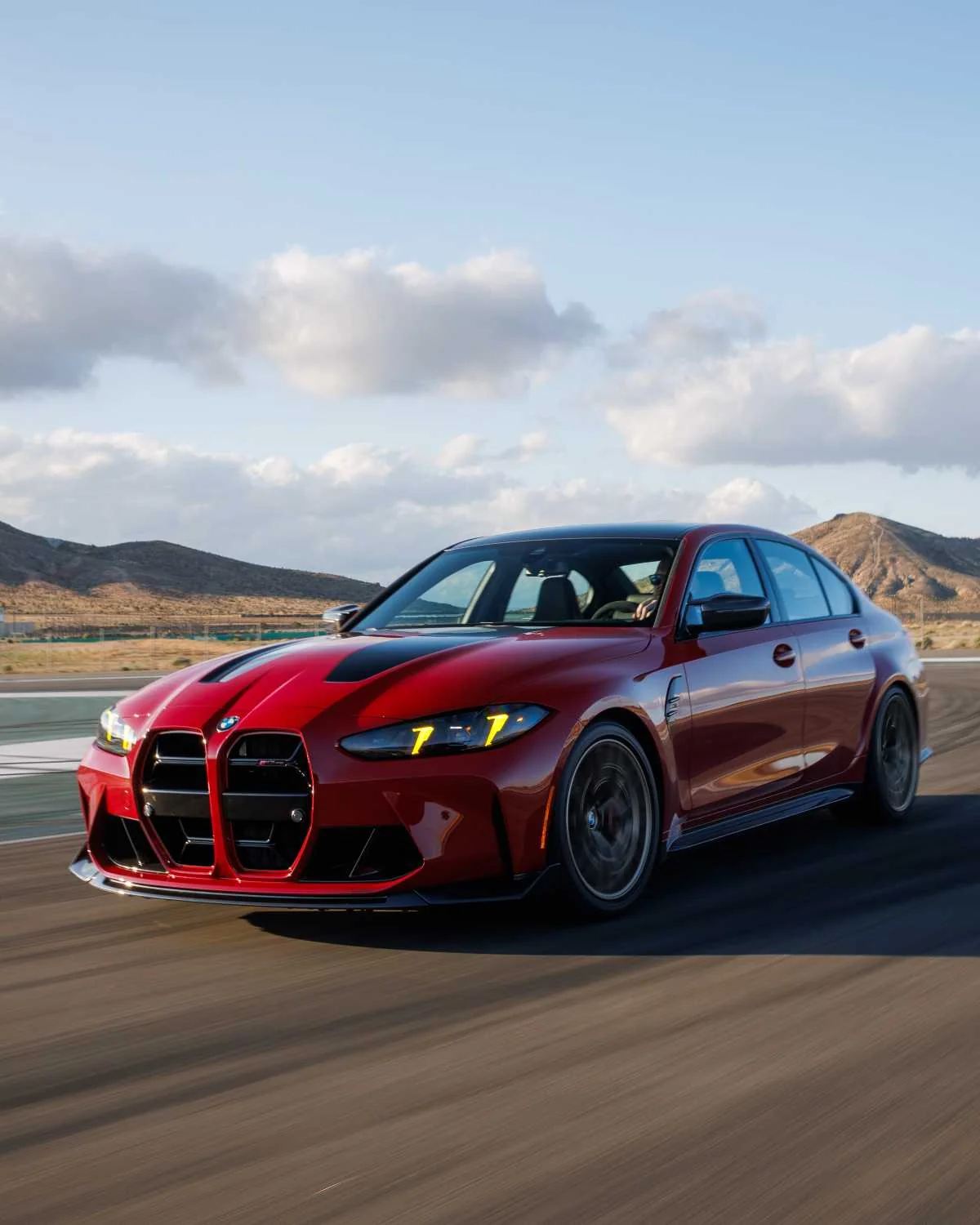 Deep red 2027 BMW M3 CS Handschalter accelerating on desert track with mountains in background under cloudy sky