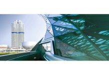 BMW Takes €700 Million Hit From Pricing Pressure as Q1 Profit Falls 25 Percent BMW vehicle parked near modern architecture with iconic tower building in background, urban setting showcasing automotive de…
