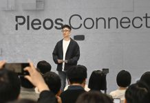 Hyundai Pleos Connect Infotainment Launches in May, Targets 20 Million Vehicles by 2030 Man presenting Hyundai Pleos Connect infotainment system at tech event, standing before gray backdrop with Pleos.Connect bra…