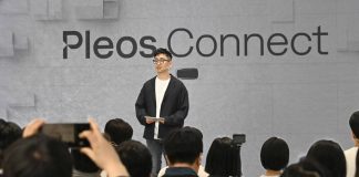 Hyundai Pleos Connect Infotainment Launches in May, Targets 20 Million Vehicles by 2030 Man presenting Hyundai Pleos Connect infotainment system at tech event, standing before gray backdrop with Pleos.Connect bra…