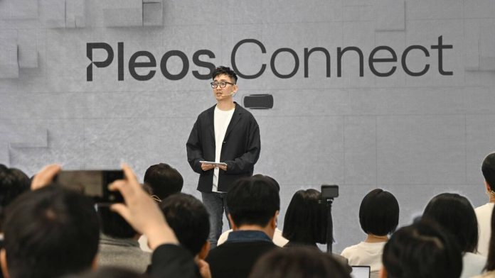 Man presenting Hyundai Pleos Connect infotainment system at tech event, standing before gray backdrop with Pleos.Connect bra…