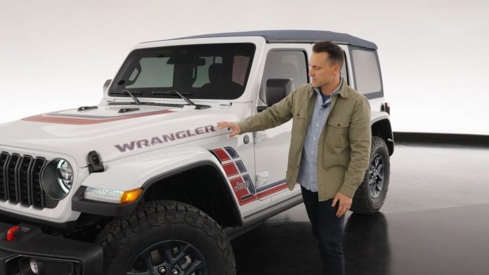 Man in tan jacket gestures toward white 2026 Jeep Wrangler America250 Edition with red and blue side graphics and exposed ho…