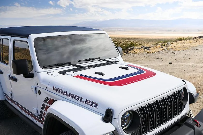 White 2026 Jeep Wrangler America250 parked in desert landscape, hood featuring red and blue Captain America shield design wi…