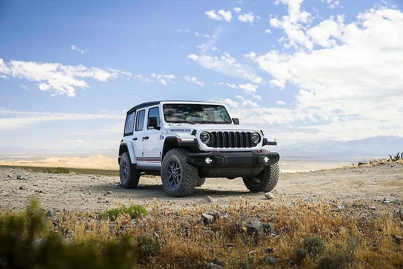 White 2026 Jeep Wrangler America250 parked on rocky desert terrain with blue sky and clouds in background.